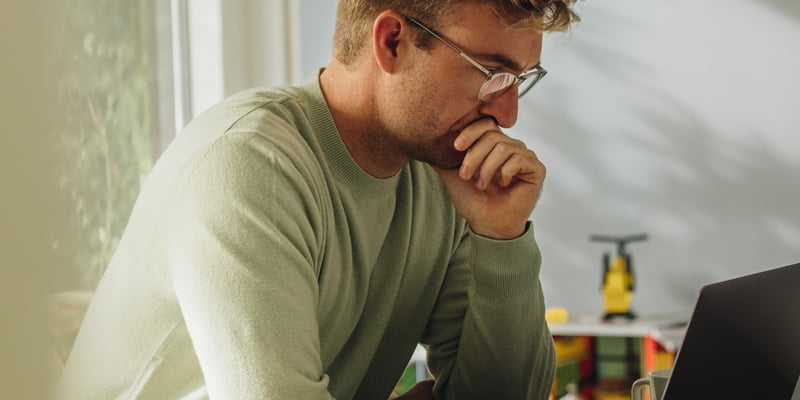 A man sitting down with his hand on his mouth looking at his laptop