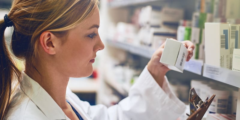 A white woman wearing a white coat, standing by a shelf with lots of medication and looking at medication in her hand.